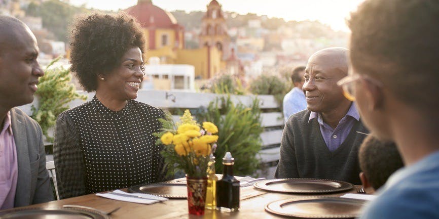 Three generations of a smiling Black family dine outdoors at sunset, the restaurant in a mountainous, warmly lit city.
