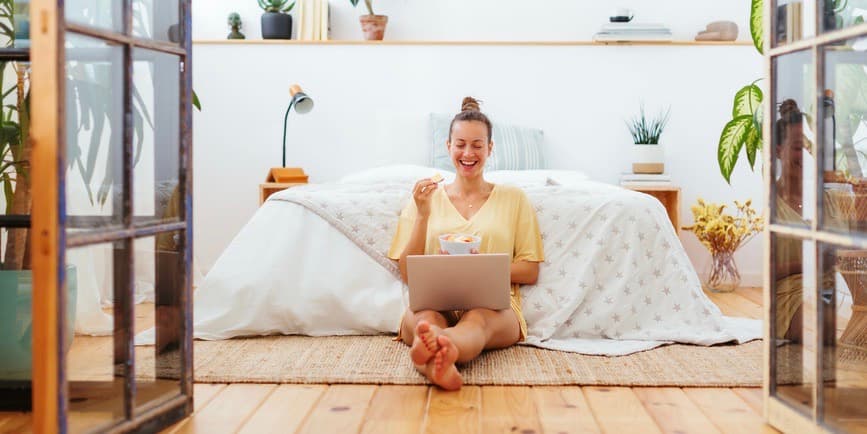 Full body image of a young white female in casual wear eating snacks and enjoying a funny movie on laptop while sitting on floor near bed in modern bedroom at home.