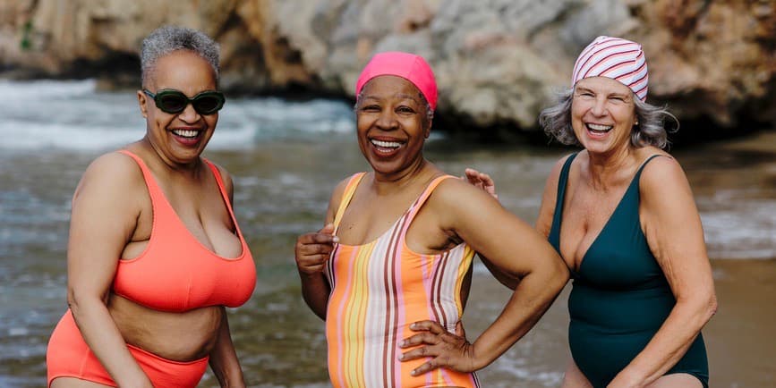 Three retired women in colorful swimsuits and bathing caps laughing and enjoying their time together on a beach vacation.