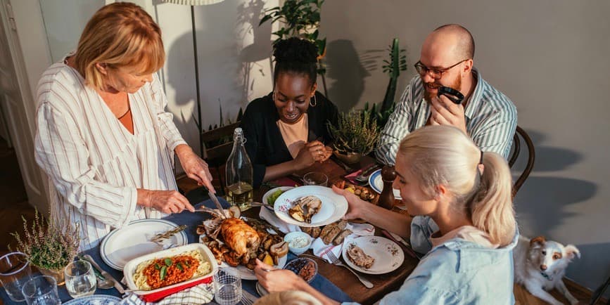 A group of friends around mid-age gather at a small table covered with food, smiling as one carves a chicken, while a small dogs rests on the floor, looking up at them.