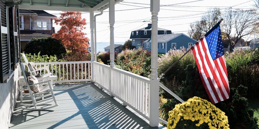 A front porch with a white fence and two white rocking chairs, an American flag blowing in the breeze, the tops of other houses and the suggestion of water in the distance.