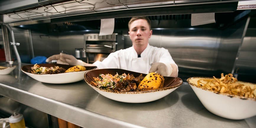 A young white cook in a commercial kitchen puts a set of finished dishes up at the pass.