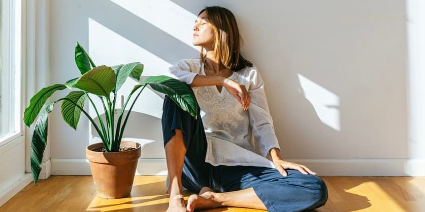 A portrait of a white woman with long blond hair wearing a white shirt and jeans, sitting on the floor in the sunlight with her eyes closed.