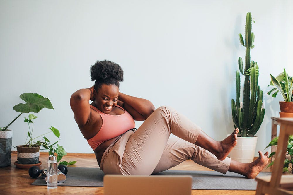 Woman working out, in a crunch position, at home. She is smiling and watching a screen.