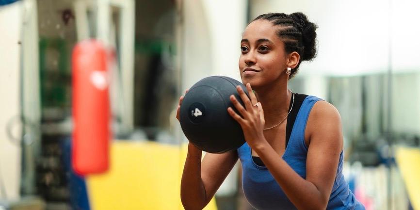 Determined Ethiopian female athlete with braided hair bun squatting with heavy medicine ball during intense workout in gym.