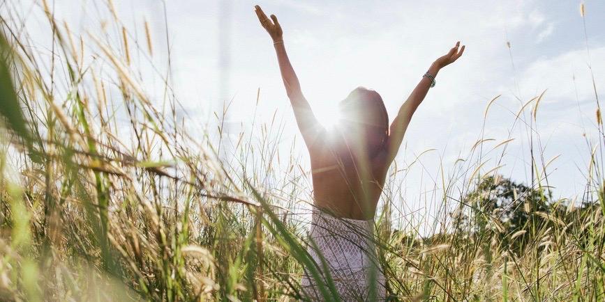 The back of a young woman standing in a field of wildflowers during sunset, her arms raised.