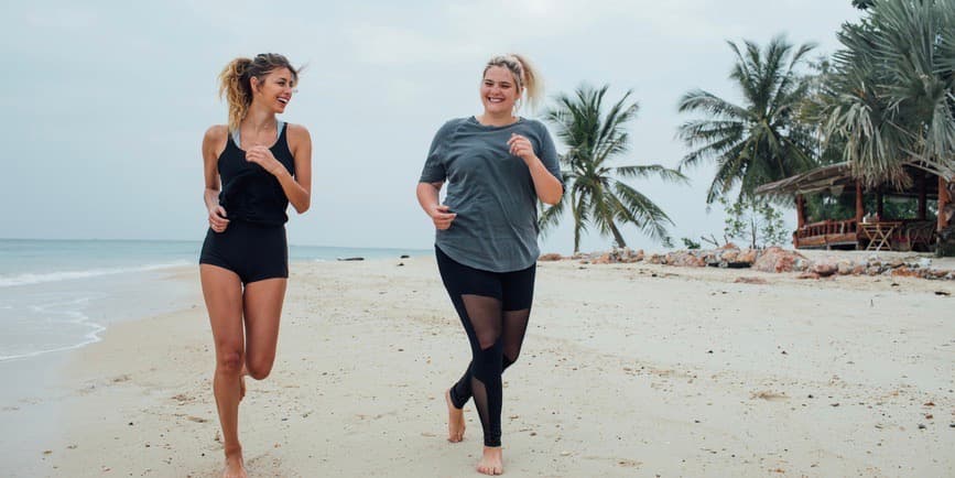 Two young white women, both with blond hair, one thin, one curvy, run on a beach in Thailand, smiling and laughing together.