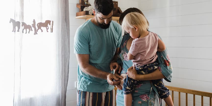 A white family with a mother holding their toddler while the father gives the mother an injection into her stomach.