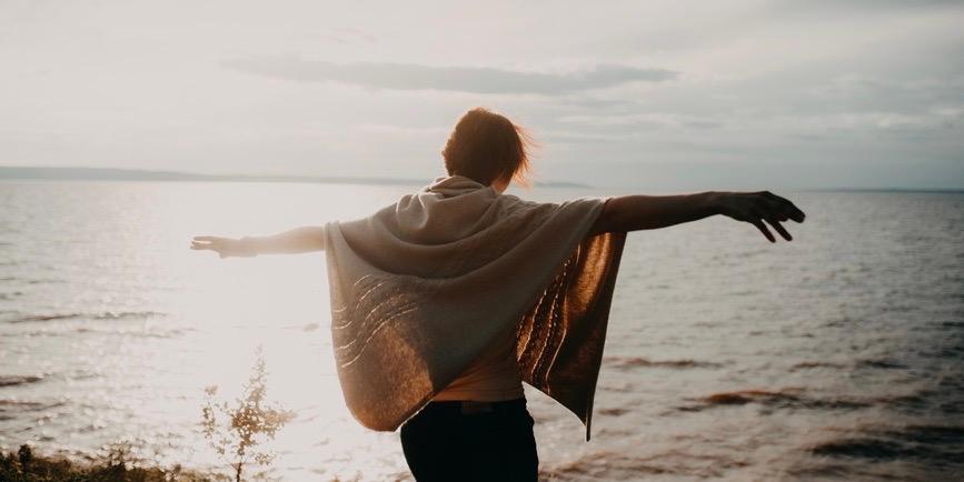 A portrait from behind of a young woman on the beach rising her hands toward the sun.