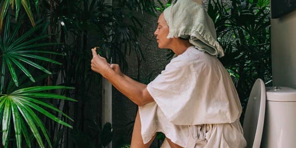 Middle-aged beautiful woman wearing beige bath robe sitting on a toilet seat looking at her phone in the outdoor bathroom.