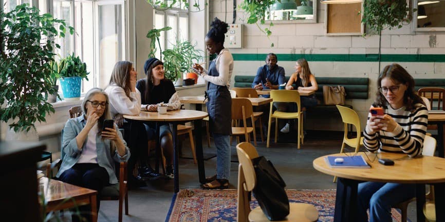 Group of multiethnic people resting and spending time together in cafe at daytime. Black waitress taking order from lesbian couple. 
