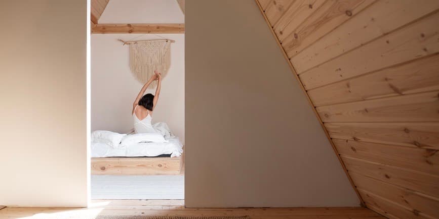 Anonymous brunette in sleepwear raising arms and stretching back while sitting on bed after awakening in sunlit attic bedroom.