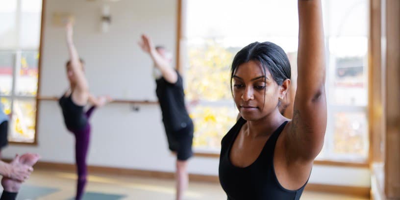 A young, thin woman with brown skin does a challenging yoga class in modern studio setting.