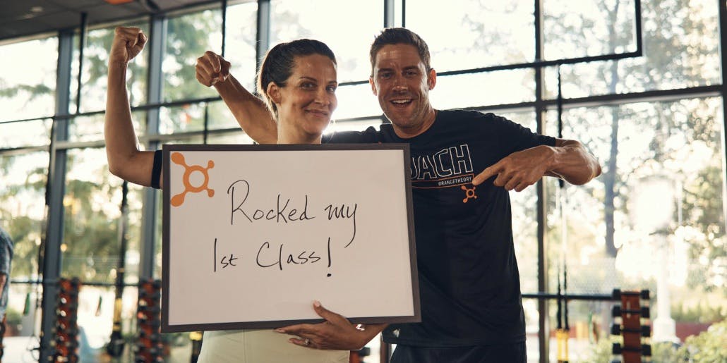 A middle-aged white woman smiles, makes a muscle and and holds a sign that says "Rocked my 1st class" while standing next to a white male Orangetheory Coach who also smiles and points down at the sign.