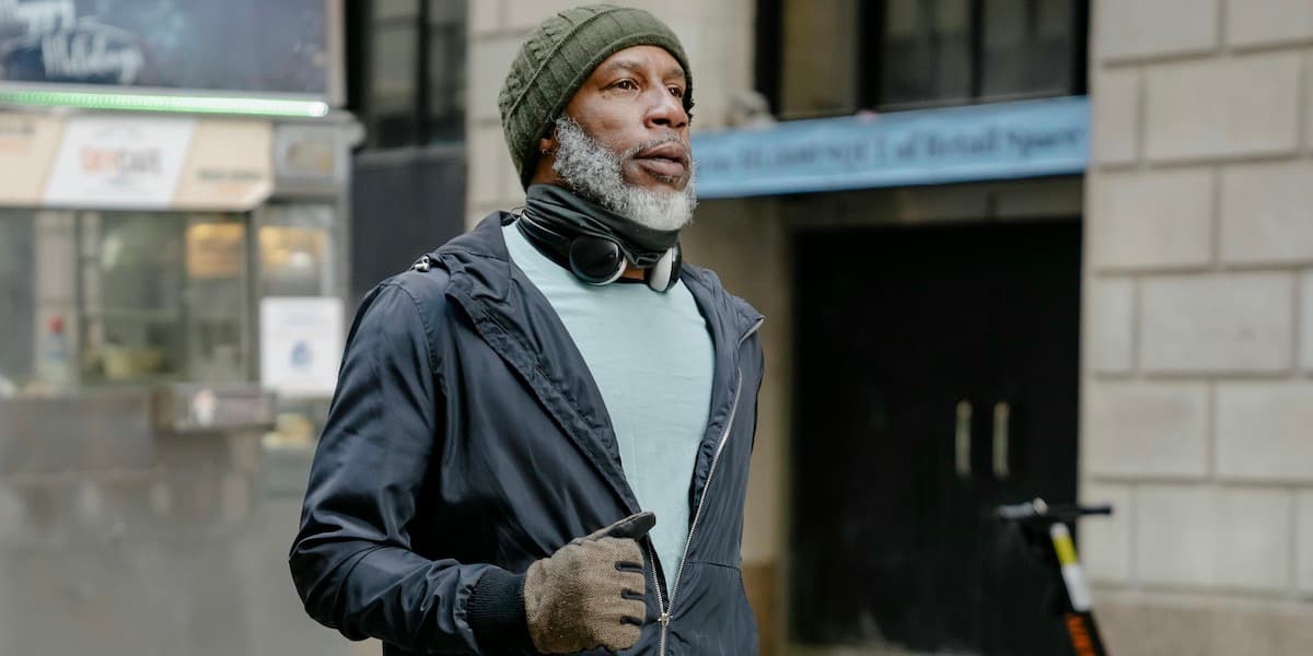 Thoughtful bearded Black man with a gray beard wearing a knit hat, gray tshirt and blue jacket jogs down a city street. 