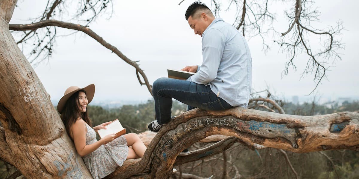 A young, attractive Asian couples sits in a tree, reading books and smiling to each other.