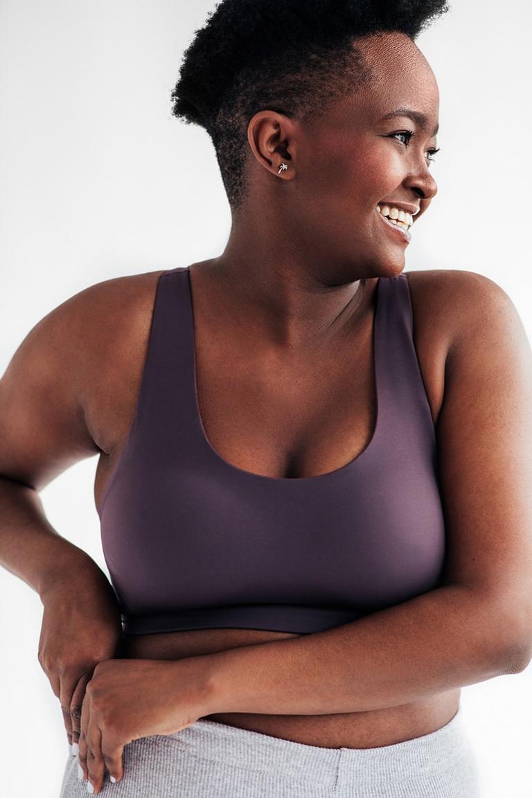 In front of a neutral background, woman in sports clothes holding right part of her waistband with both hands, looking and smiling to the left.