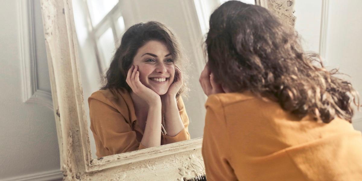 A young woman with long brown hair wearing an orange shirt smiles as she rests her head in her hands and looks at herself in a mirror.