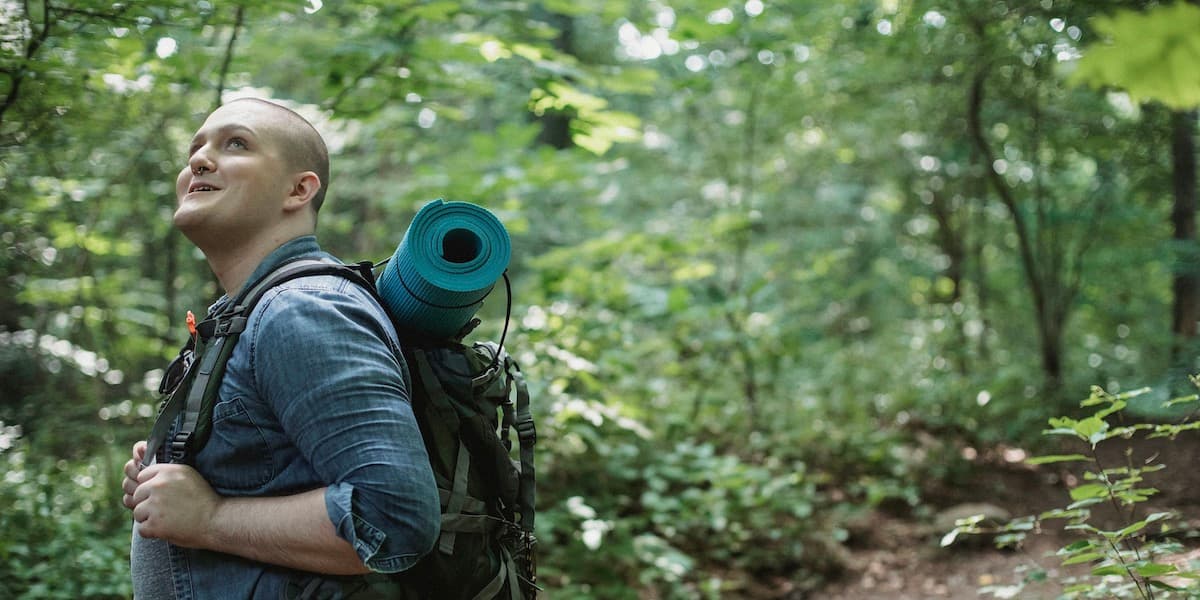 A male hiker with no hair wearing a blue shirt and a backpack with a yoga mat on top smiles as he looks up at something in the trees.