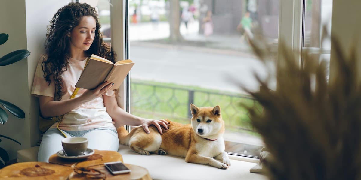 A white woman with long dark curly hair sits in the window of a coffee shop reading a book and petting her shiba inu dog, who lays in the window seat beside her.