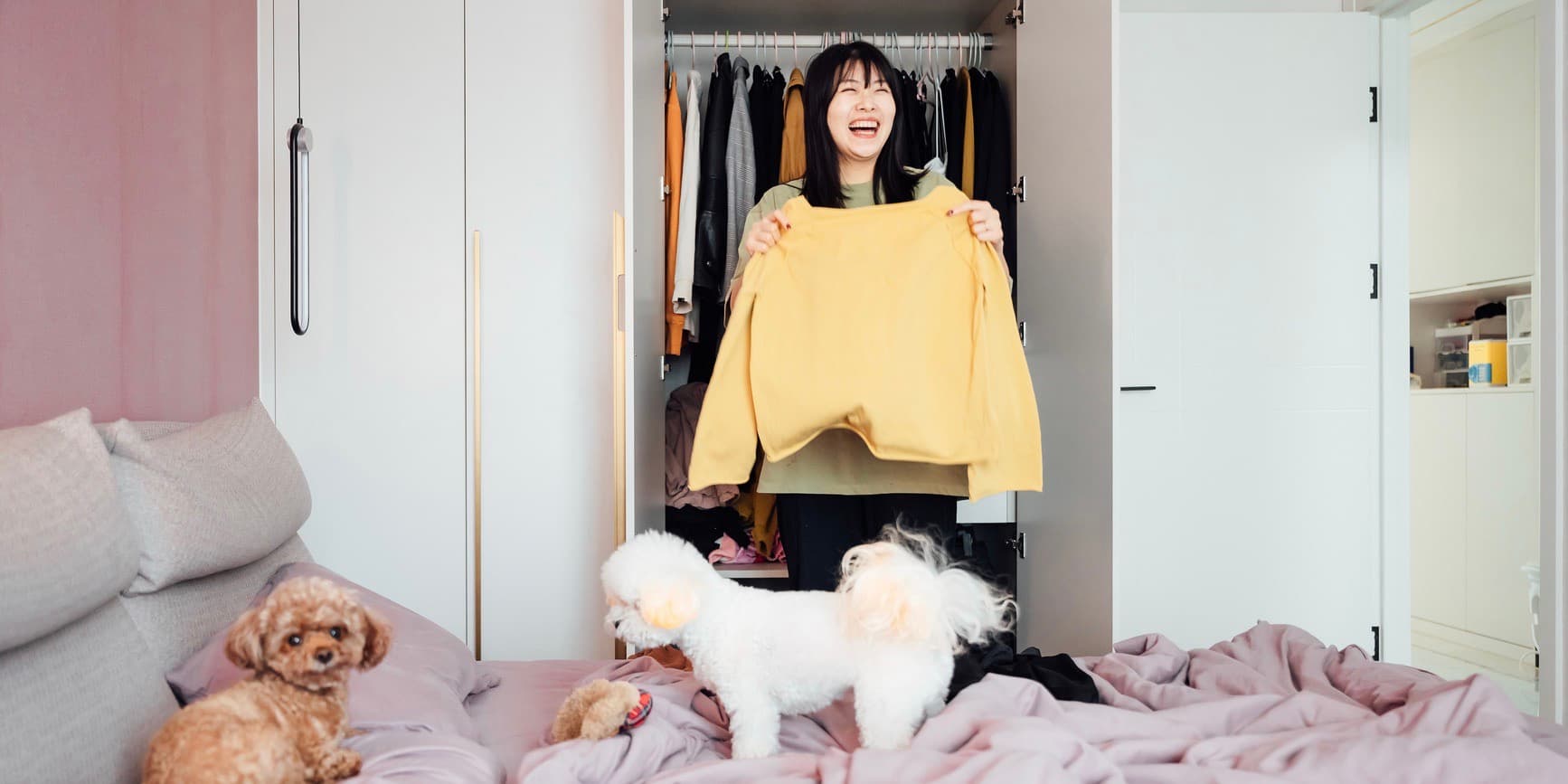 Young Asian woman smiling as she holds a sweater up in front of her closet, choosing what to wear, with her two fluffy dogs on her bed.