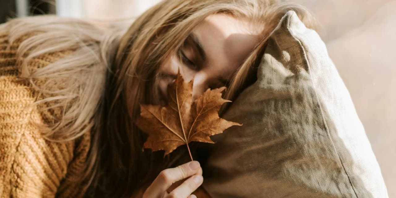 Closeup shot of a young white woman with long dark blonde hair wearing an orange sweater cuddling on a pillow and holding a maple leaf close to her face with a cup of tea nearby.