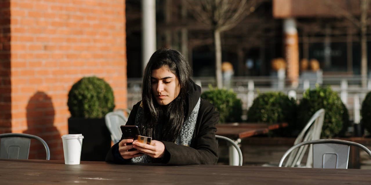 A young woman with dark skin and long dark hair wearing a black winter coat sits outside at a cafe table in the sun, looking down into her phone with a credit card out to pay for something.
