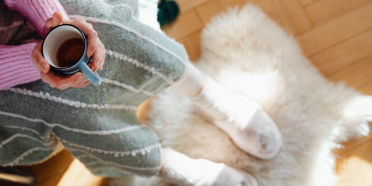 Overhead shot of a woman's sweatered arms holding a mug of coffee or tea in her hands, which are resting on a blanket, and her slippered feet, which rest on a white sheepskin rug.