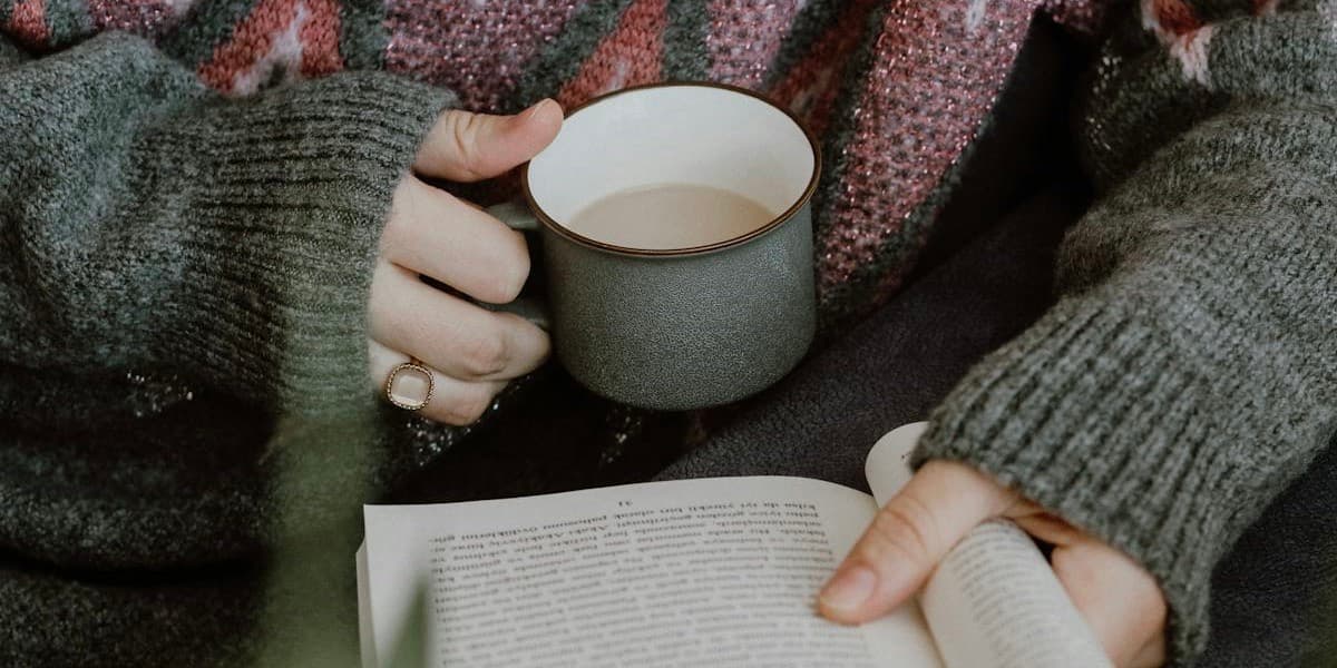 A closeup of a white woman's heavily sweatered hands holding a mug of coffee and a book.