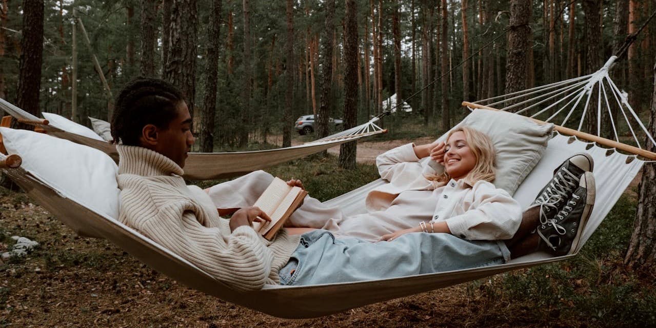 A young Black man and young white woman in stylish fall clothes lie in a hammock in the middle of the woods, reading and smiling at each other.