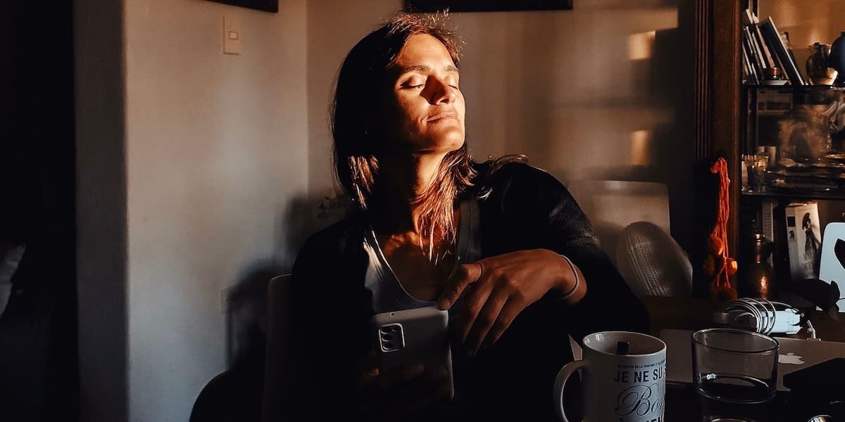 A middle aged white woman with brown hair sits at a table in a cluttered dining room with a mug and a phone, her face lit by morning sunlight, smiling with her eyes shut.