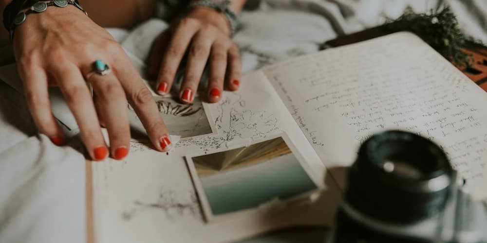 A closeup shot of a white woman's hands with funky jewelry and chipped red nail polish over a journal with photos and sketches, and a camera nearby.