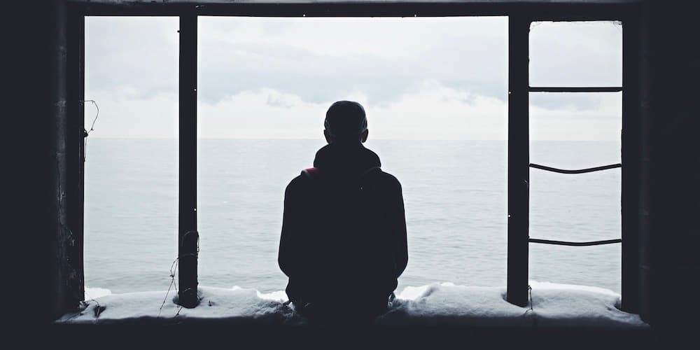 A photo of a human in shadow, taken from behind. The person is sitting in an open window covered in snow with their legs outside, looking out into a stark white landscape.