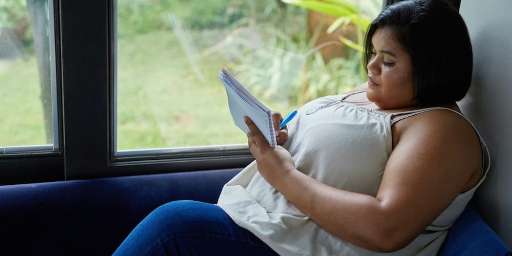 An attractive woman with a heavier weight, brown skin and short black hair wearing a tank top and jeans writes in a journal while sitting on a blue velvet couch next to a large window with greenery visible outside.