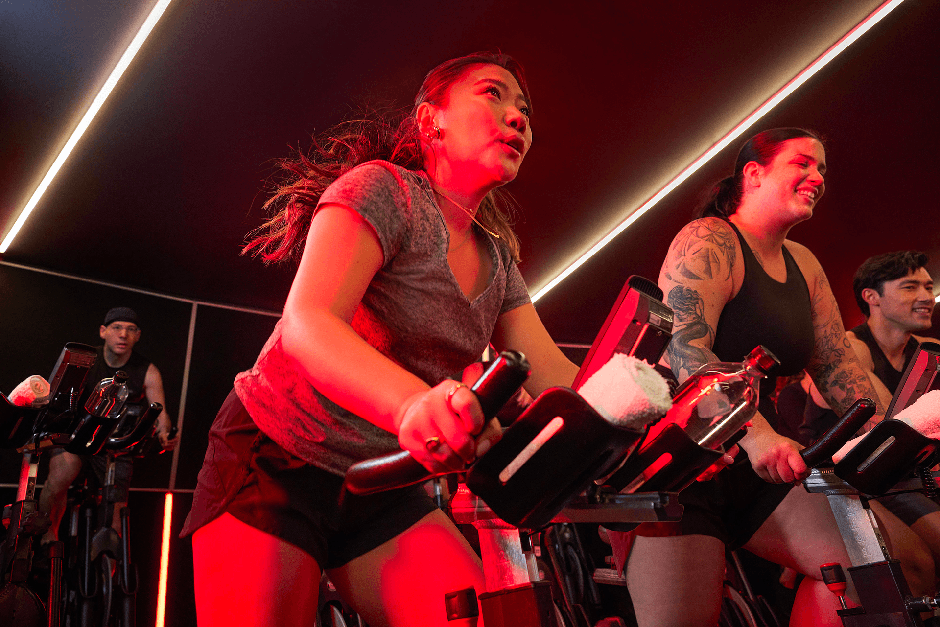 A group of people in a dimly lit indoor cycling class, pedaling intensely under red lighting. A central woman in a gray t-shirt leads the effort, while others, including a smiling tattooed woman, ride alongside her.