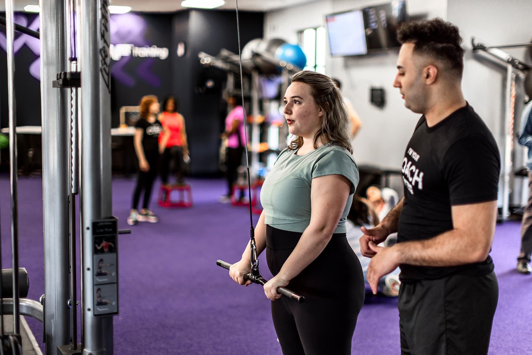 A fitness coach provides one-on-one guidance to a member using a cable machine in a vibrant gym setting with others training in the background.