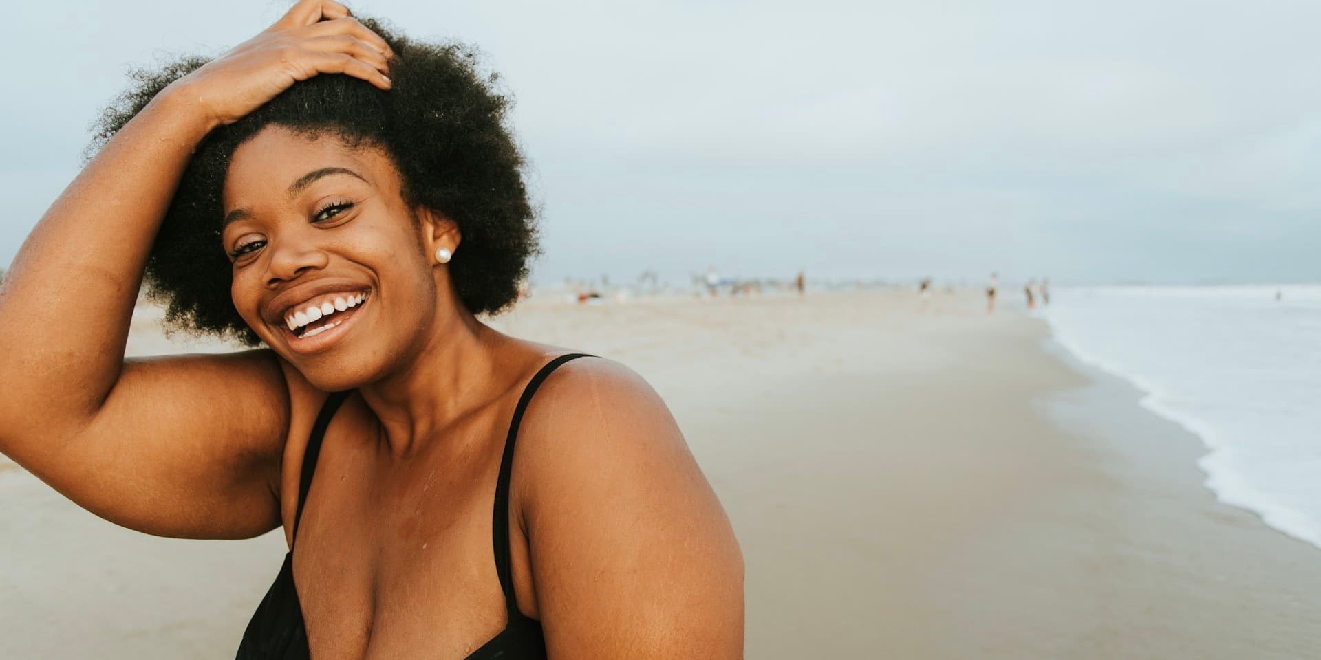 A curvy Black woman wearing a black bathing suit stands on a beach and smiles wide into the camera as she runs her fingers through her hair.