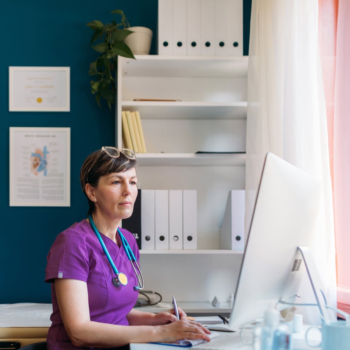 A medical professional wearing purple scrubs working at her desk on a computer in her office