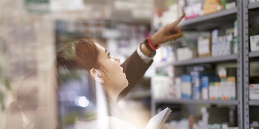 A pharmacist points to shelves of medication inside of a pharmacy to another pharmacist with a notebook