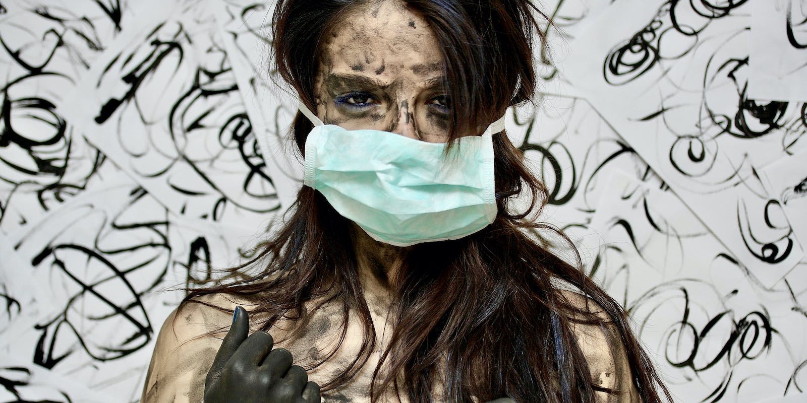 A masked, exhausted woman covered in mud stares intensely at the camera against a backdrop of muddy, artistic swirls on a white wall