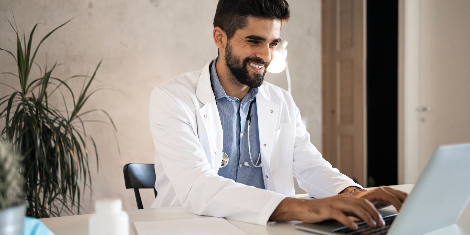 A male doctor with medium-toned skin and dark hair sits in a blue shirt and lab coat with a stethoscope around his neck, typing into a computer and smiling