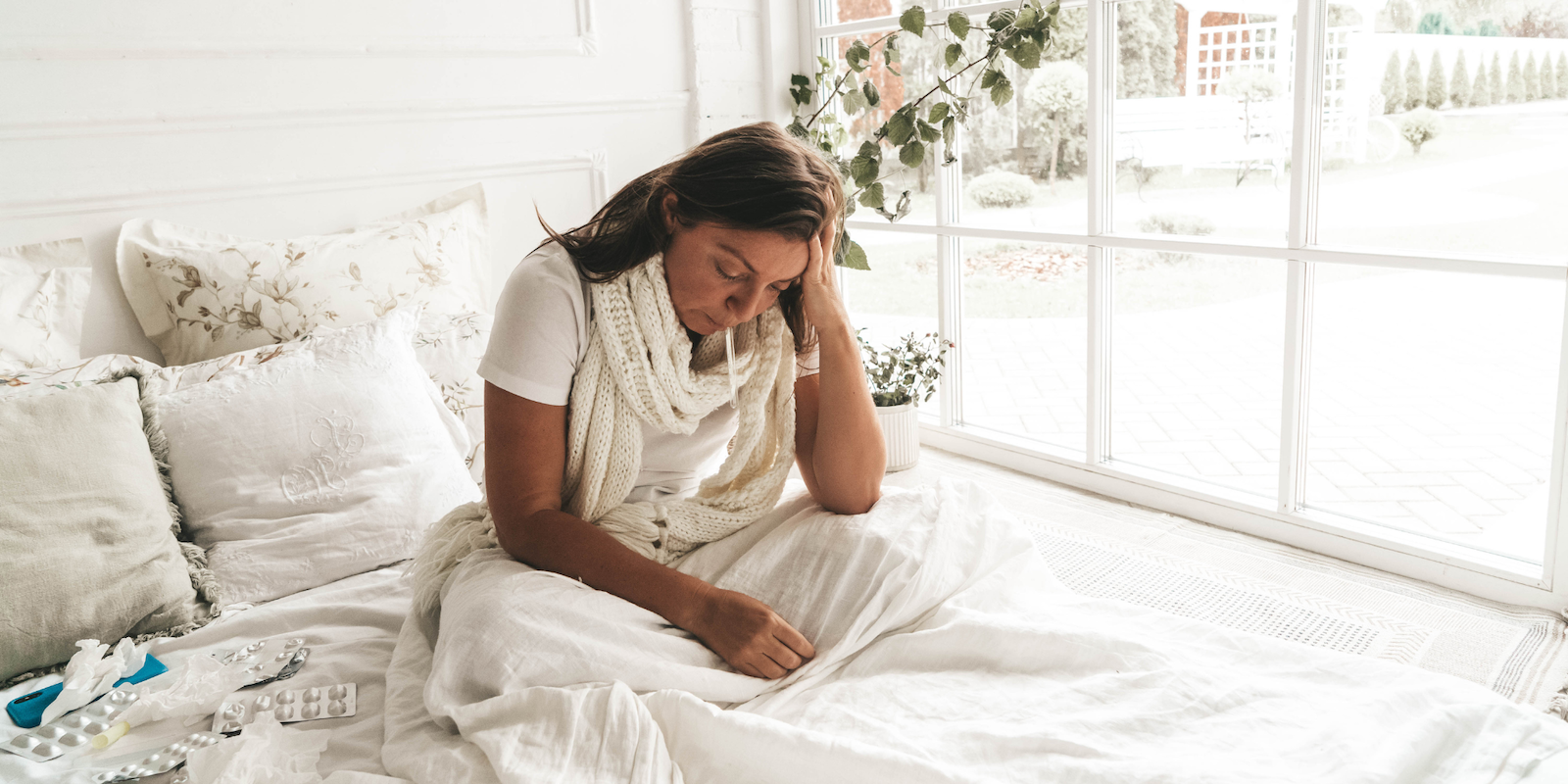 Fotografía de una mujer enferma sentada en la cama con la cabeza entre las manos, rodeada de medicamentos.