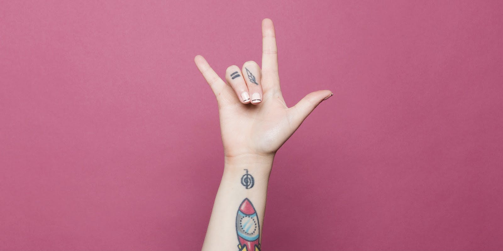 A tattooed forearm reaches up against a bright pink background with a hand spelling "I love you" in sign language.