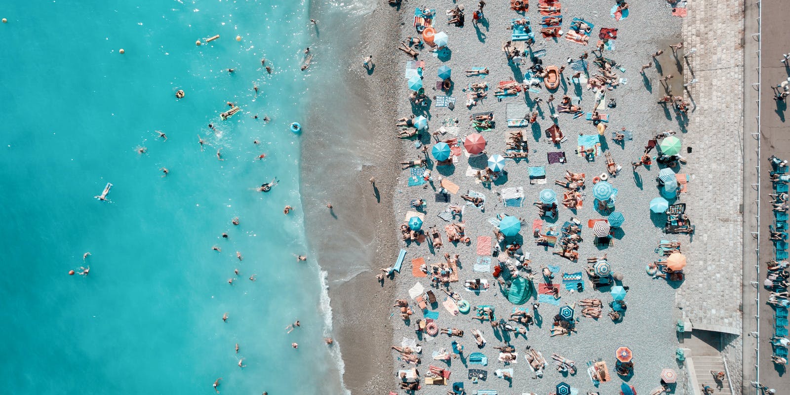 A high overhead photograph of a Mediterranean beach with turquoise water to the left and sand dotted with umbrellas and people on the right.