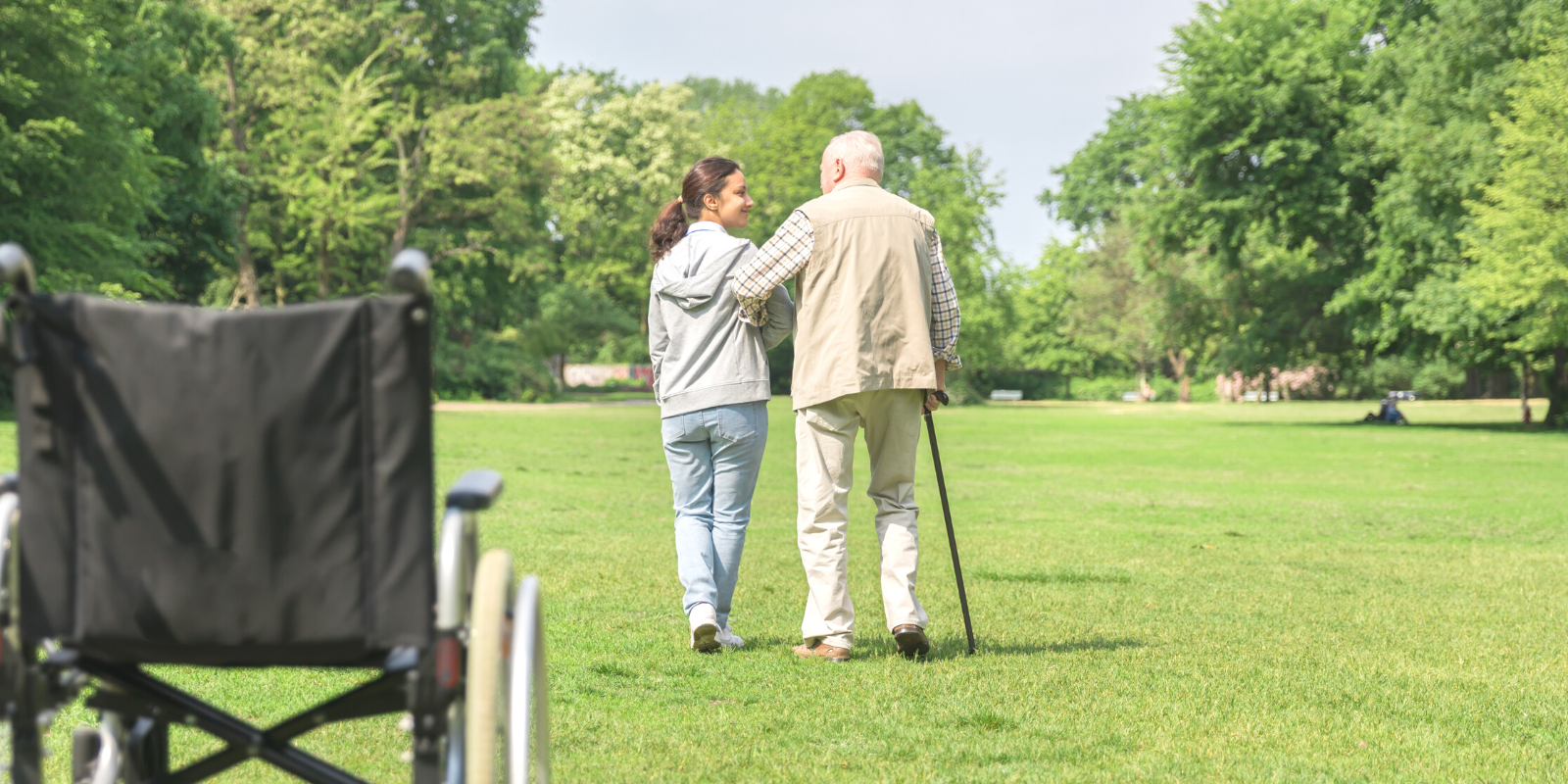 A photograph of a large green field, with an older white man with gray hair walking with a cane and a younger woman with brown skin and hair wearing jeans and a sweatshirt, both walking away from the camera, with an empty wheelchair in the foreground.