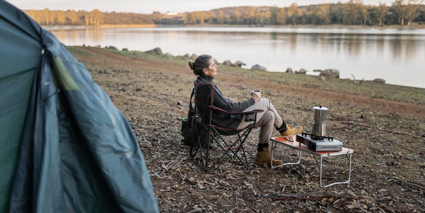 A photograph of a white woman in her fifties sitting in a camp chair on a pebbled beach, looking into the water with a table and a coffee pot next to her, and a tent almost out of the frame.