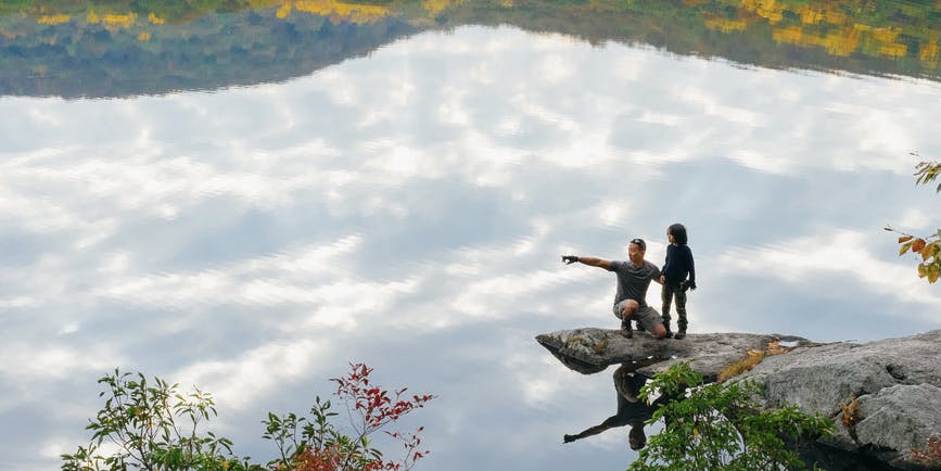 A color photograph of a wide outdoor autumn landscape. And Asian adult man and young teenage child stand on a rock cliff high over a body of water, which reflects the clouds and autumnal trees at top and bottom.