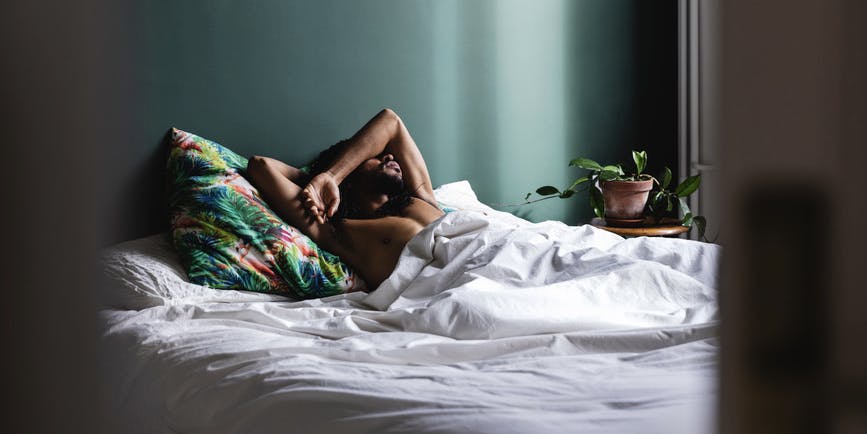 A color photograph of a Black man sleeping in a bed with white sheets and colorful green and orange pillow, in a bed set against a solid deep green wall, their arm shielding their eyes from daylight.