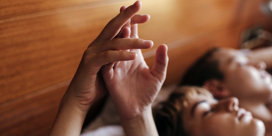 A color photograph of two white people laying in bed against a wooden headboard, their hands intertwined in the air.