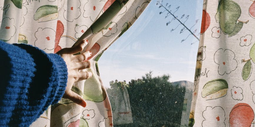 A color photograph of a window with an arm in a blue sweater pulling back faded curtains with fruit on them, a blue sky showing outside.